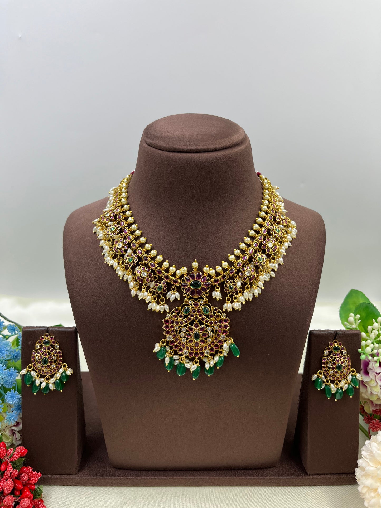 Gold necklace with green and red accents on a brown mannequin bust, surrounded by decorative flowers.